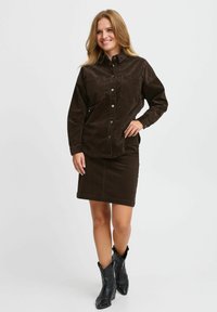 Camicia di velluto a coste marrone con chiusura a bottoni e due tasche sul petto, abbinata a una gonna di velluto a coste marrone coordinata. Stivali cowboy neri completano l'outfit.