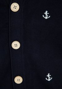 Námořnický pletený kardigan s dřevěnými knoflíky a bílou výšivkou kotvy. Má texturovanou látku a přední otvor na celou délku.