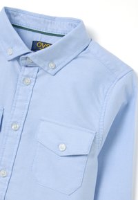 Camicia blu chiaro in cotone misto, con colletto, due tasche sul petto e bottoni bianchi. Texture liscia.