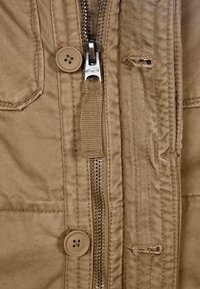 Beżowa kurtka z metalowym zamkiem błyskawicznym i dwoma dużymi guzikami, z widocznymi przeszyciami i fakturowaną tkaniną. Zawiera naszywaną kieszeń.
