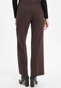 Pantalons marron à jambes larges avec une texture lisse, dotés de deux poches arrière et d'un design de taille épuré. Le modèle porte des bottes à talons noires.