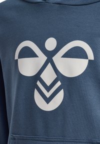 Felpa con cappuccio di colore blu scuro realizzata in tessuto morbido, caratterizzata da un grande motivo grafico bianco che assomiglia a un insetto stilizzato con forme geometriche.