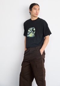 Czarny bawełniany t-shirt z kolorowym wzorem graficznym, na którym znajdują się zabawne ikony oraz napis "NAJLEPSZY W KLASIE", zestawiony z brązowymi spodniami cargo.