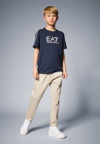 Jūrų stiliaus marškiniai su "EA7 EMPORIO ARMANI" grafika, kreminės spalvos kelnės ir baltos sportbačiai. Ant marškiniai yra kontrastingos šoninės juostelės.