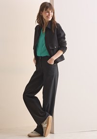 Giacca nera con tasche frontali, blusa teal e pantaloni neri a gamba larga, abbinati a sneakers chiare con design bianco e beige.