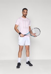 Αθλητική μπλούζα σε ανοιχτό ροζ χρώμα με τυπωμένο σχέδιο, λευκό σορτς, μαύρα παπούτσια τένις και μαύρο βραχιολάκι καρπού· κρατώντας ρακέτα τένις.