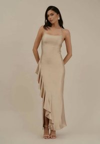 Vestido longo de cetim bege com corpete ajustado e uma barra assimétrica com pregas. Apresenta alças finas e uma fenda lateral. Textura suave.