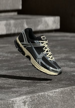 Černá a šedá běžecká obuv Nike s krémovými tkaničkami vznášející se nad texturovaným betonovým povrchem.