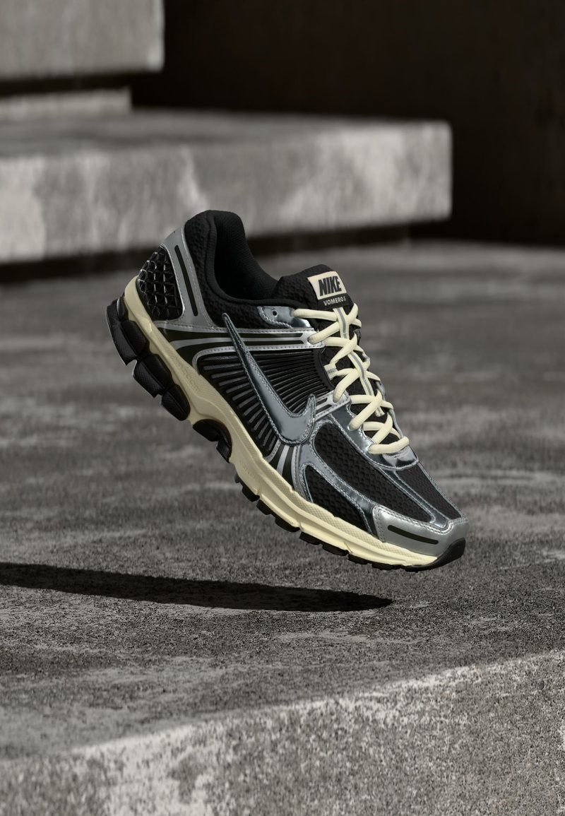 Chaussure de course Nike noire et grise avec des lacets crème flottant au-dessus d'une surface en béton texturée.