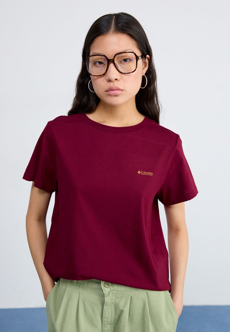 Burgundowa koszulka z krótkim rękawem, okrągłym dekoltem i małym złotym logo. Noszona z jasnoniebieskimi spodniami o wysokim stanie.