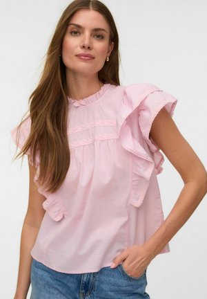 Kvinde iført en lyserød bluse med flæseærmer og blonde detaljer, kombineret med blå jeans, poserer med den ene hånd i lommen.
