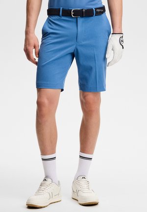 Mann trägt blaue Golfshorts, schwarzen Gürtel, weißen Golfhandschuh, weiße Socken mit schwarzen Streifen und weiße Sneakers vor einfarbigem Hintergrund.