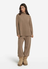 Brauner Kabelstrick-Pullover mit hohem Kragen, kombiniert mit passenden, lässig geschnittenen Hosen und flauschigen, braunen Stiefeln. Weiche Textur über das gesamte Outfit.