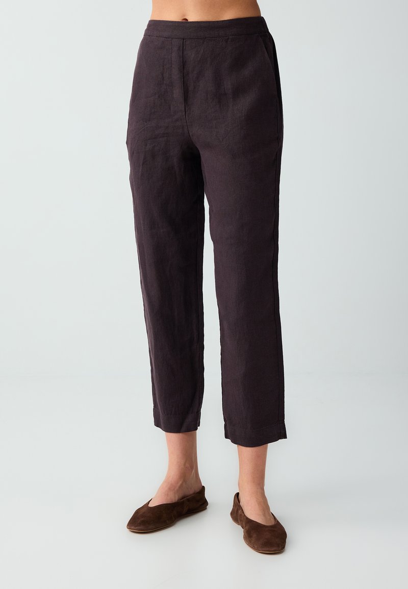 Femme portant un pantalon court marron foncé et des chaussures marron à enfiler assorties, debout sur un fond gris clair.