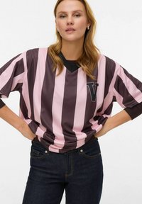 Perdidelis marškinėlis su tamsiai rudos ir šviesiai rožinės spalvos vertikaliomis juostomis, juodas V formos kaklas ir „V“ logotipas kairėje krūtinės pusėje.