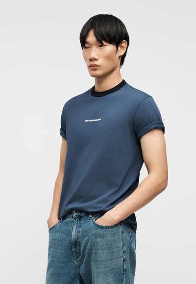 Νεαρός άνδρας με μαύρα μαλλιά φοράει μπλε μπλουζάκι Emporio Armani με ανεβάσματα στα μανίκια και ανοιχτόχρωμο τζιν, στέκεται με τα χέρια στις τσέπες.