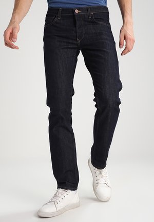 Mand iført mørkeblå slim fit jeans og hvide sneakers, stående mod en ensfarvet lys baggrund.