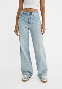 Ljust blå vida jeans i denim med hög midja, femfickorsdesign och knappstängning. Bärs med silverfärgade sneakers.