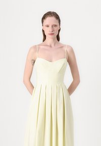 Rochie plisată de un galben deschis, cu decolteu în formă de inimă și bretele subțiri. Textura materialului este netedă, cu un corset ajustat și un design cu fustă evazată.