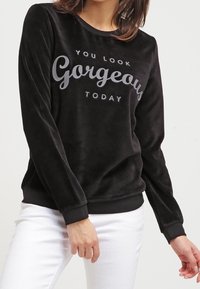 Sudadera de terciopelo negra con mangas largas, puños y dobladillo acanalados. Presenta el texto impreso "TE VES HERMOSA HOY" en gris. Combinada con pantalones blancos.