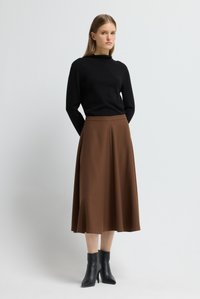 Frau in schwarzem langärmligem Oberteil, brauem Midikleid und schwarzen Stiefeletten, die vor einem einfarbigen hellen Hintergrund steht.