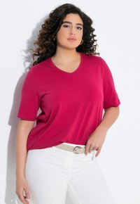 T-shirt à manches courtes et col en V en tissu magenta vif, coupe légèrement ample, associé à un jean blanc et une ceinture de couleur claire.