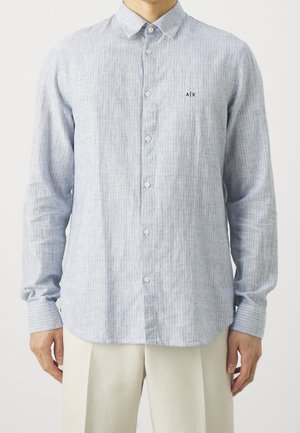 Muž nosící světle modrou košili s dlouhými rukávy na knoflíky s jemnými vertikálními pruhy a malým logem A|X na levé straně hrudi, v kombinaci s béžovými kalhotami.
