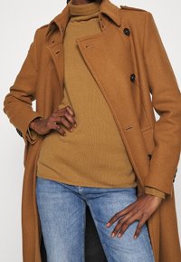 Brązowy wełniany dwurzędowy płaszcz z szerokim kołnierzem, zestawiony z beżowym prążkowanym golfem i niebieskimi dżinsami, prezentowany na białym tle.