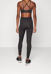 Černá sportovní podprsenka s křížovými ramínky, v kombinaci s černými legínami s výřezovými akcenty na kotnících a texturovanou tkaninou. Součástí jsou také sportovní boty.