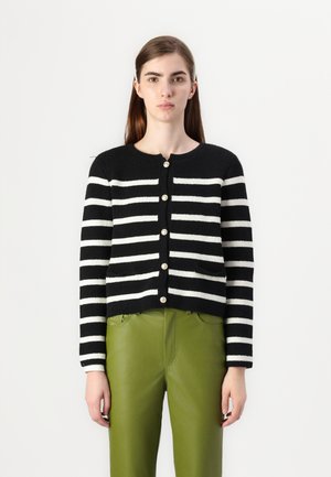 Černo-bílý pruhovaný cardigan s kulatým výstřihem, zdobený zlatými knoflíky a dvěma předními kapsami, nošený přes zelené kožené kalhoty.