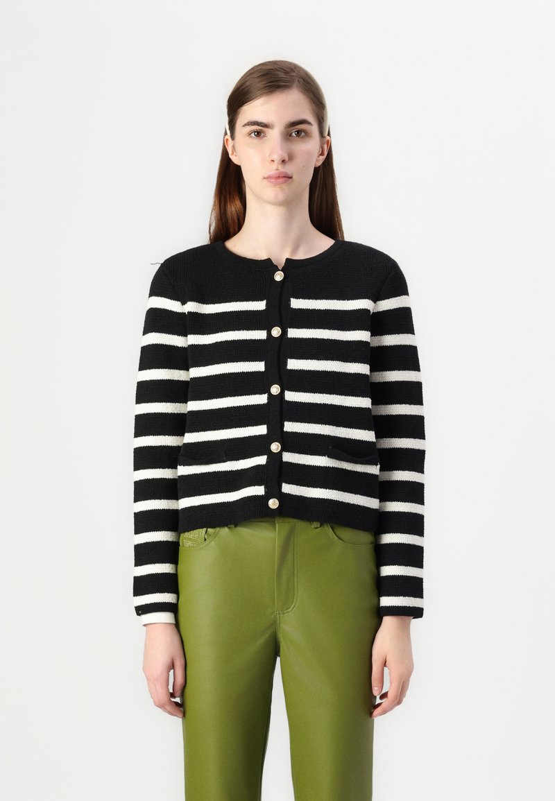 Cardigan a righe bianche e nere con scollatura rotonda, dotato di bottoni dorati e due tasche frontali, indossato sopra pantaloni di pelle verde.