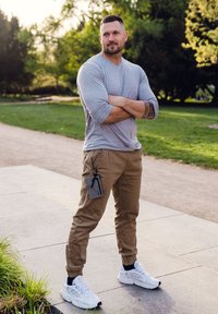 Gray long-sleeve shirt and tan jogger pants with cuffed hems; white sneakers. Phone attached to pants via a black accessory. Green park background.