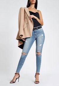 Blazer oversize color beige claro con botones dorados, top de satén negro, jeans ajustados de mezclilla azul claro desgastados y tacones negros con tiras y detalles de estampado de leopardo.
