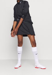 Musta pitkähihainen urheilu-toppi tekstuuripinnalla, yhdistetty mustan hameen kanssa. Valkoiset sukat ja punaisilla yksityiskohdilla varustetut kengät täydentävät asun.