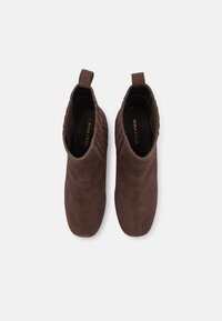 Botas de tornozelo em camurça castanha com uma textura suave, apresentando puxadores laterais e um design de bico arredondado. Acentuadas com detalhes de costura sutis.