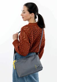 Borsa a tracolla grigia con motivo e finitura texture, accentata da una cerniera gialla e con tracolla regolabile, indossata sopra un maglione di colore ruggine.