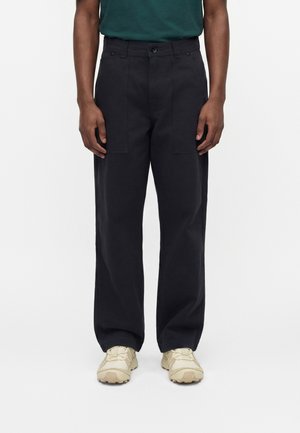 Άνδρας με μαύρο παντελόνι ίσιας γραμμής με μεγάλες μπροστινές τσέπες, μπεζ αθλητικά παπούτσια και πράσινο πουκάμισο, στέκεται μπροστά σε απλό λευκό φόντο.