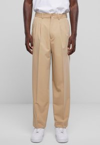 Beige op maat gemaakte broek met geplooide voorkant, gemaakt van een gladde stof, met een knoopsluiting en elastische tailleband. Draag het met witte sneakers.