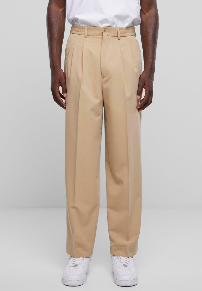 Beige op maat gemaakte broek met geplooide voorkant, gemaakt van een gladde stof, met een knoopsluiting en elastische tailleband. Draag het met witte sneakers.