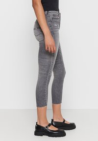 Jeans skinny de mezclilla gris con dobladillo deshilachado y detalles desgastados, combinados con zapatos Mary Jane de plataforma negros. Vista lateral.