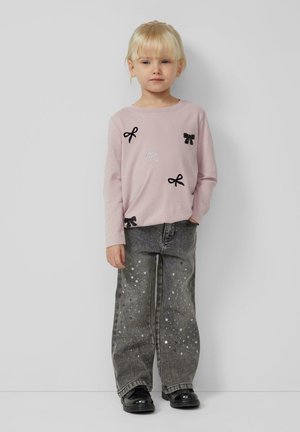 Hellrosa langärmliges Hemd mit schwarzen und weißen Schleifenapplikationen, kombiniert mit grauen Jeans mit silbernen Pailletten. Schwarze Schuhe vervollständigen das Outfit.