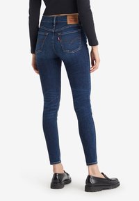 Persona vistiendo unos jeans ajustados de color azul oscuro y mocasines negros, de pie con la espalda hacia la cámara sobre un fondo blanco liso.