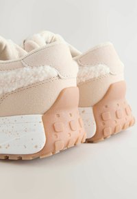 Baskets beiges avec un dos texturé, présentant un accent en peluche et une semelle intermédiaire blanche avec des motifs mouchetés. Semelle extérieure en gomme épaisse.