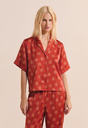 Donna bionda che indossa un completo pigiama in raso rosso con motivi gialli a forma di sole, top a maniche corte con bottoni e pantaloni larghi abbinati, in piedi davanti a uno sfondo beige.