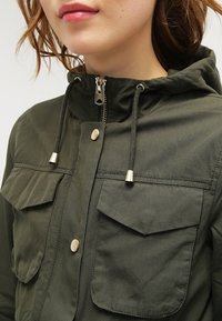 Oliwkowo-zielona kurtka z kapturem, zapinana na zamek, z regulowanymi sznurkami i dwiema klapowymi kieszeniami z metalowymi ozdobami. Wykonana z teksturowanej tkaniny.