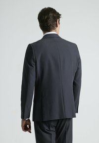Chaqueta de traje ajustada en gris oscuro, con una abertura en la parte trasera, hombros estructurados, dos botones en las mangas, textura suave, vista trasera sobre un fondo claro.