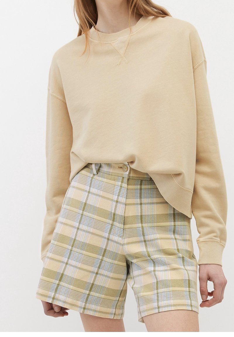 Sudadera beige con hombros caídos combinada con pantalones cortos de cuadros amarillos claros que presentan detalles en verde y azul, con cierre de botón visible.