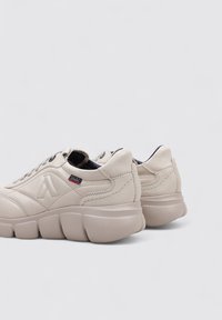 Zapatillas de cuero beige con una suela gruesa, que presentan un diseño acolchado y un acento oscuro en el talón. Incluyen una etiqueta de marca cerca del cordón.