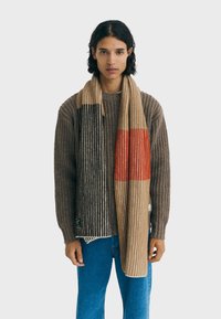 Knit scarf featuring vertical stripes in beige, brown, and orange. Soft texture with distinct color blocks and fringed edges.