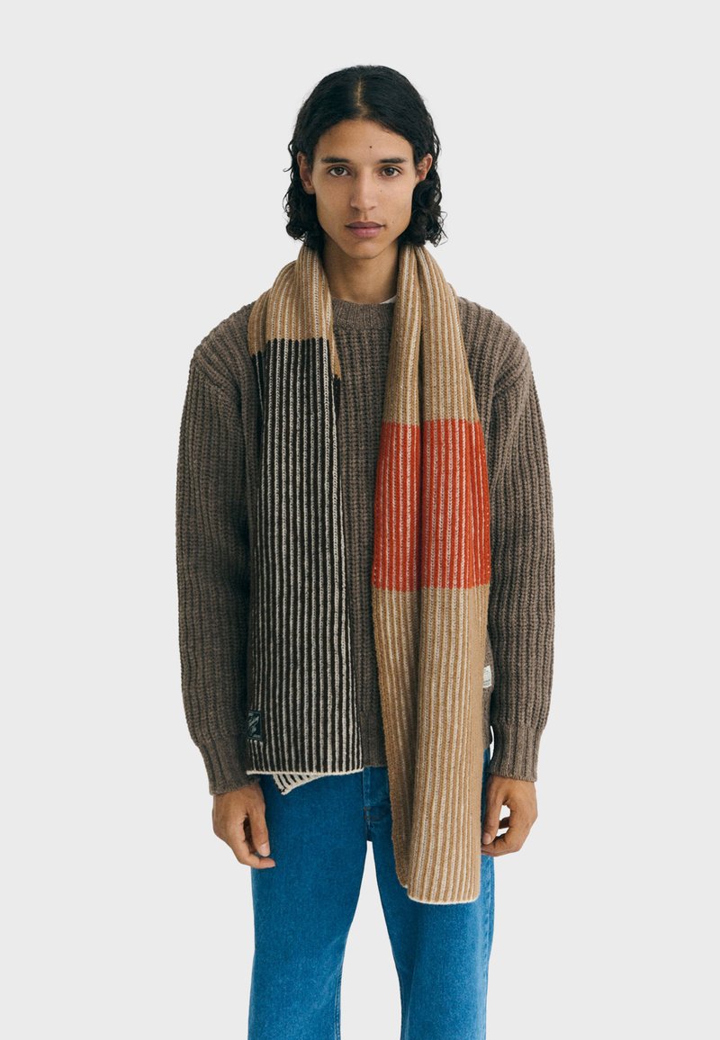 Knit scarf featuring vertical stripes in beige, brown, and orange. Soft texture with distinct color blocks and fringed edges.